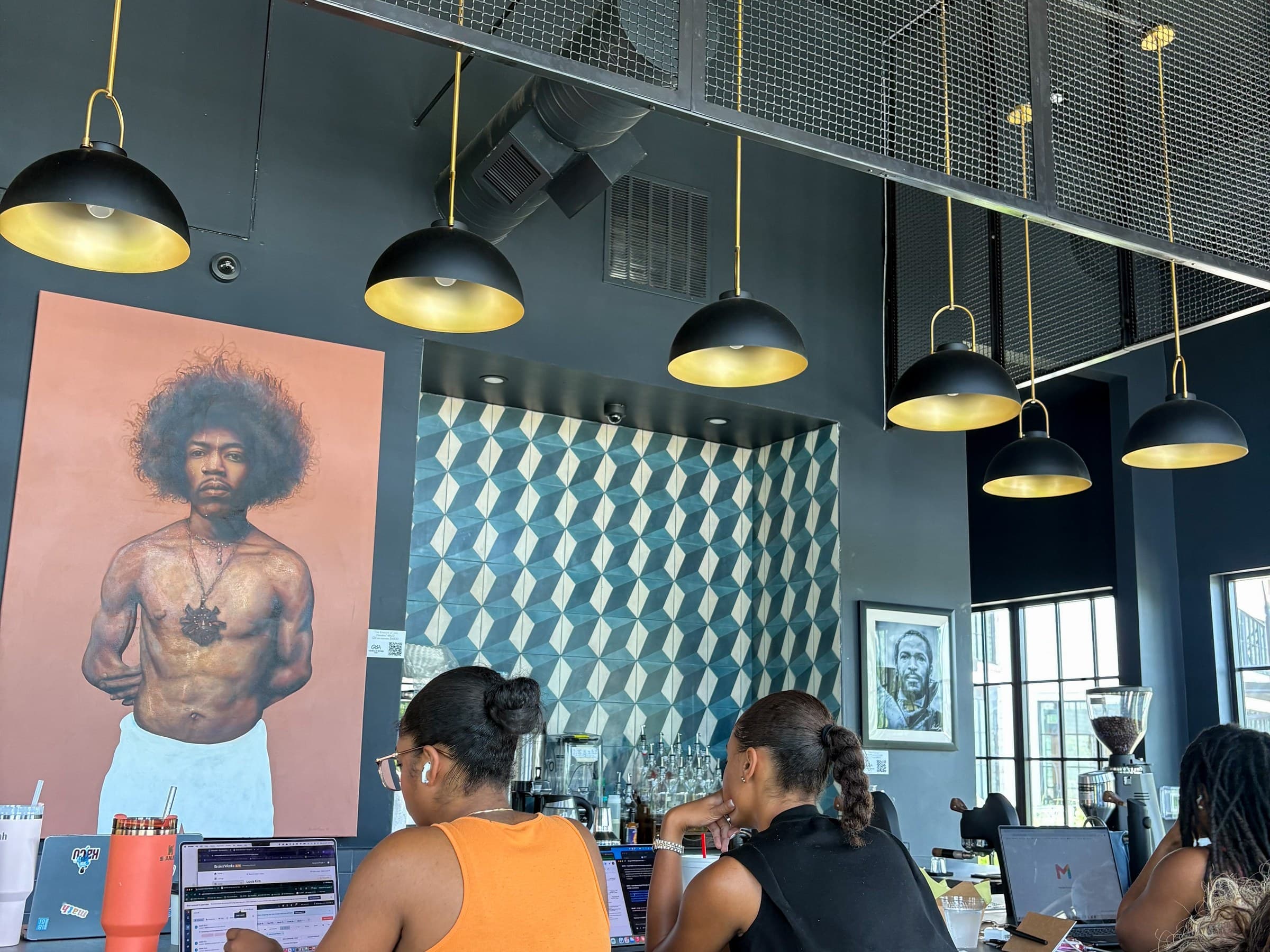 Hanging pendant lights and a geometric tiled wall above two people working on laptops at a coffee shop counter.
